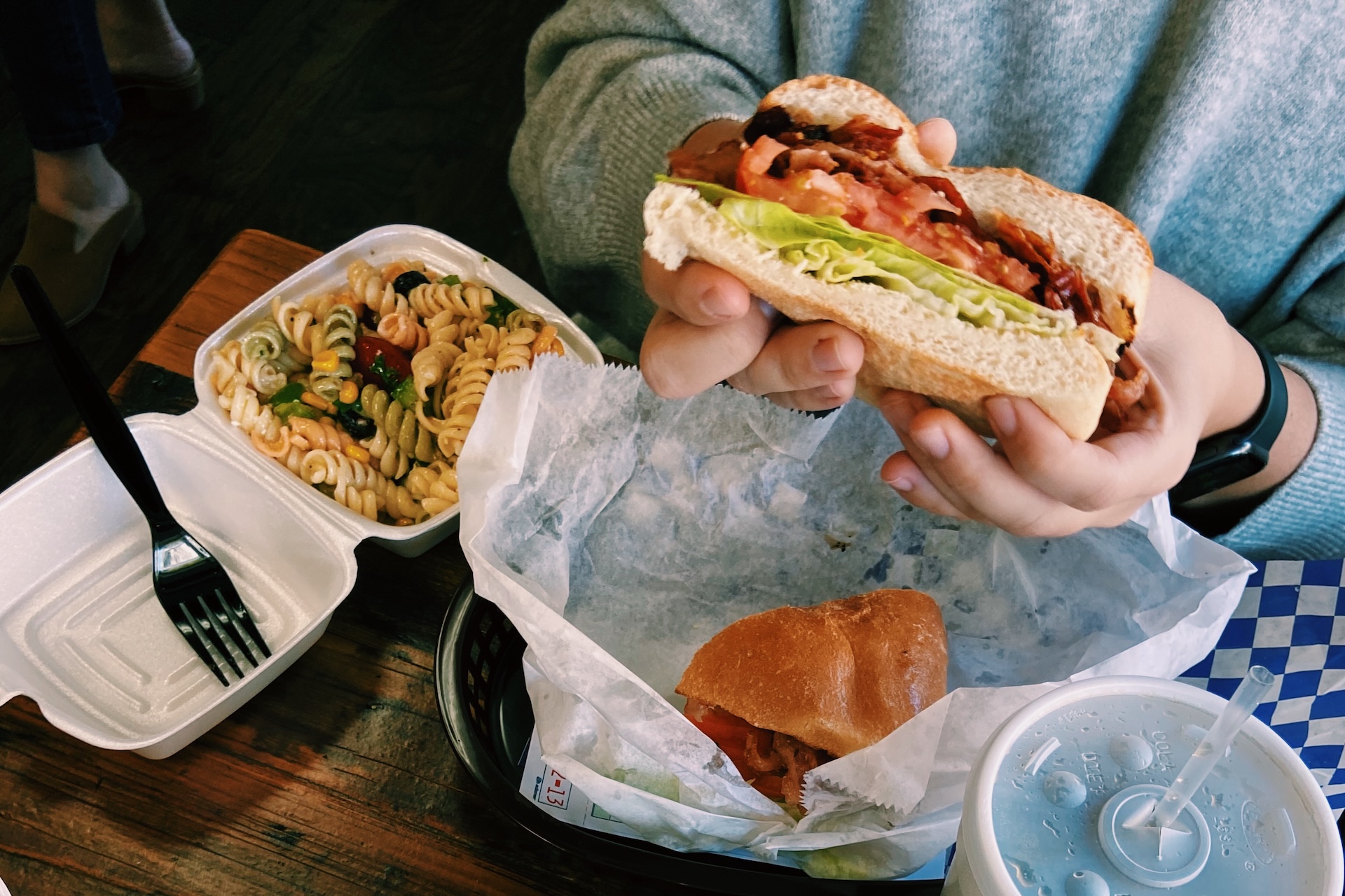 A person in a gray sweater holds a sandwich with lettuce, tomato, and bacon. On the table are a pasta salad in a takeout container, another sandwich, and a drink with a straw.