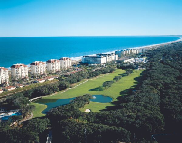 Aerial view of a coastal resort area with tall beachfront buildings, a golf course with a pond, and the ocean stretching along the horizon under a clear blue sky.