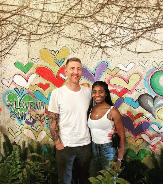 A man and woman smile while standing together in front of a colorful mural filled with overlapping hearts and vine-covered walls. Both wear casual outfits; the man has a white shirt and gray pants, and the woman has a white tank top and jeans.