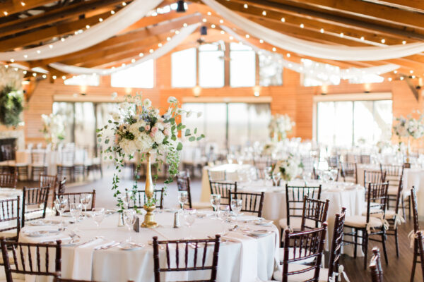 Elegant wedding reception hall with round tables covered in white linens, floral centerpieces, wooden chairs, draped fabric, and string lights hanging from a wooden-beamed ceiling.