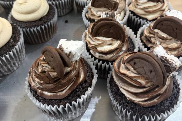Chocolate cupcakes with swirled chocolate and vanilla frosting, topped with chocolate wafer pieces and chunks of marshmallow, arranged on a metal tray.