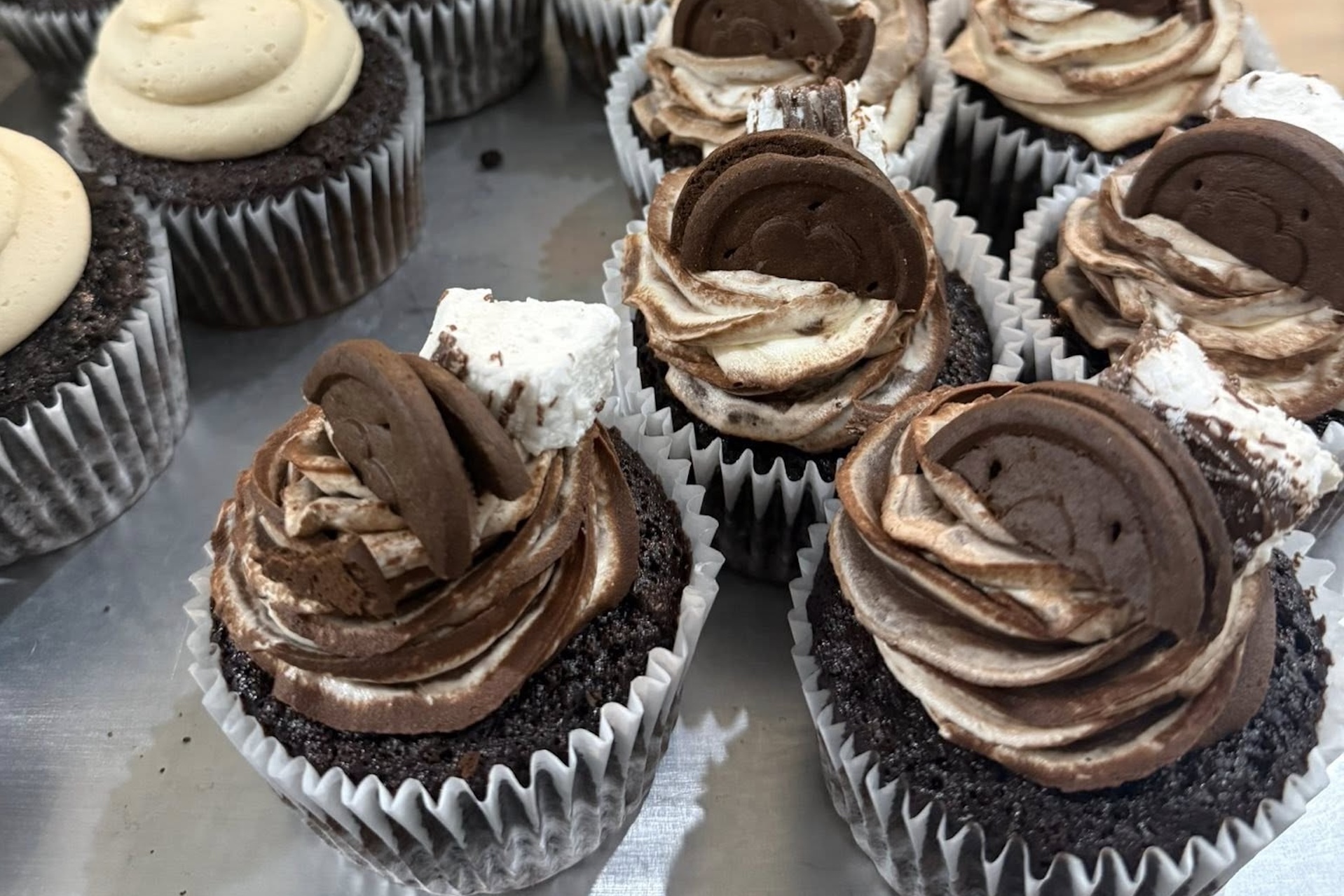 Chocolate cupcakes with swirled chocolate and vanilla frosting, topped with chocolate wafer pieces and chunks of marshmallow, arranged on a metal tray.