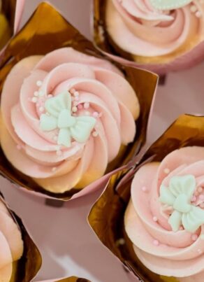 Cupcakes with pink swirled frosting, decorated with white butterfly-shaped toppers and small white sprinkles, are arranged in gold cupcake wrappers.