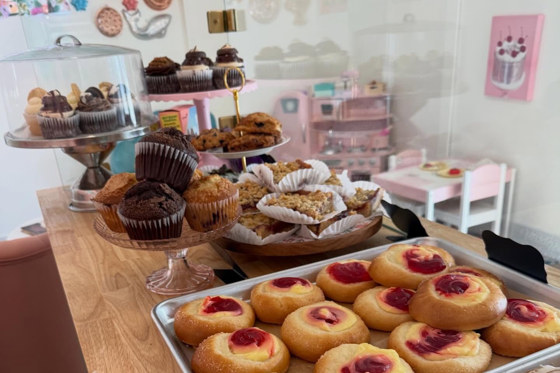 An assortment of pastries and cupcakes displayed on trays and cake stands in a bakery, with muffins, cookies, crumb cakes, and fruit-filled danishes on a wooden counter in a bright, pastel-themed setting.