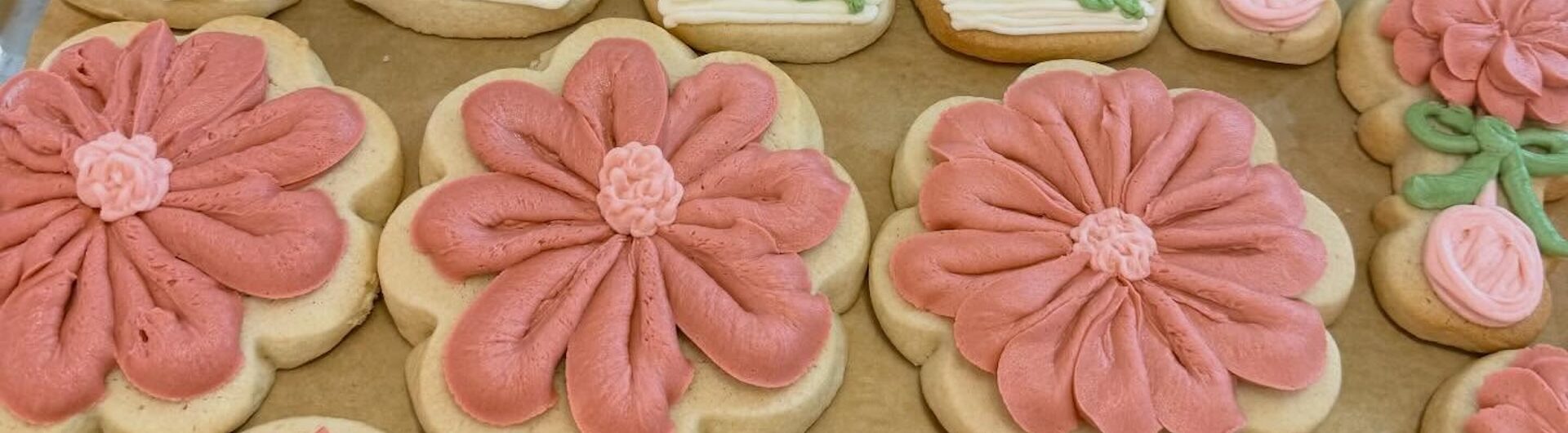 A tray of decorated sugar cookies shaped like baby bottles and flowers, with pink, white, and green icing. Some cookies are large flowers, while others feature small flowers and greenery details.