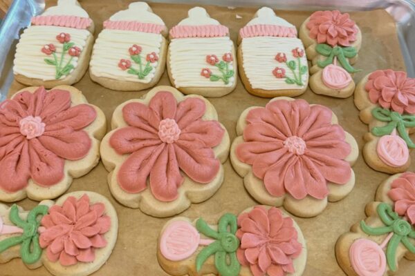 A tray of decorated sugar cookies shaped like baby bottles and flowers, with pink, white, and green icing. Some cookies are large flowers, while others feature small flowers and greenery details.