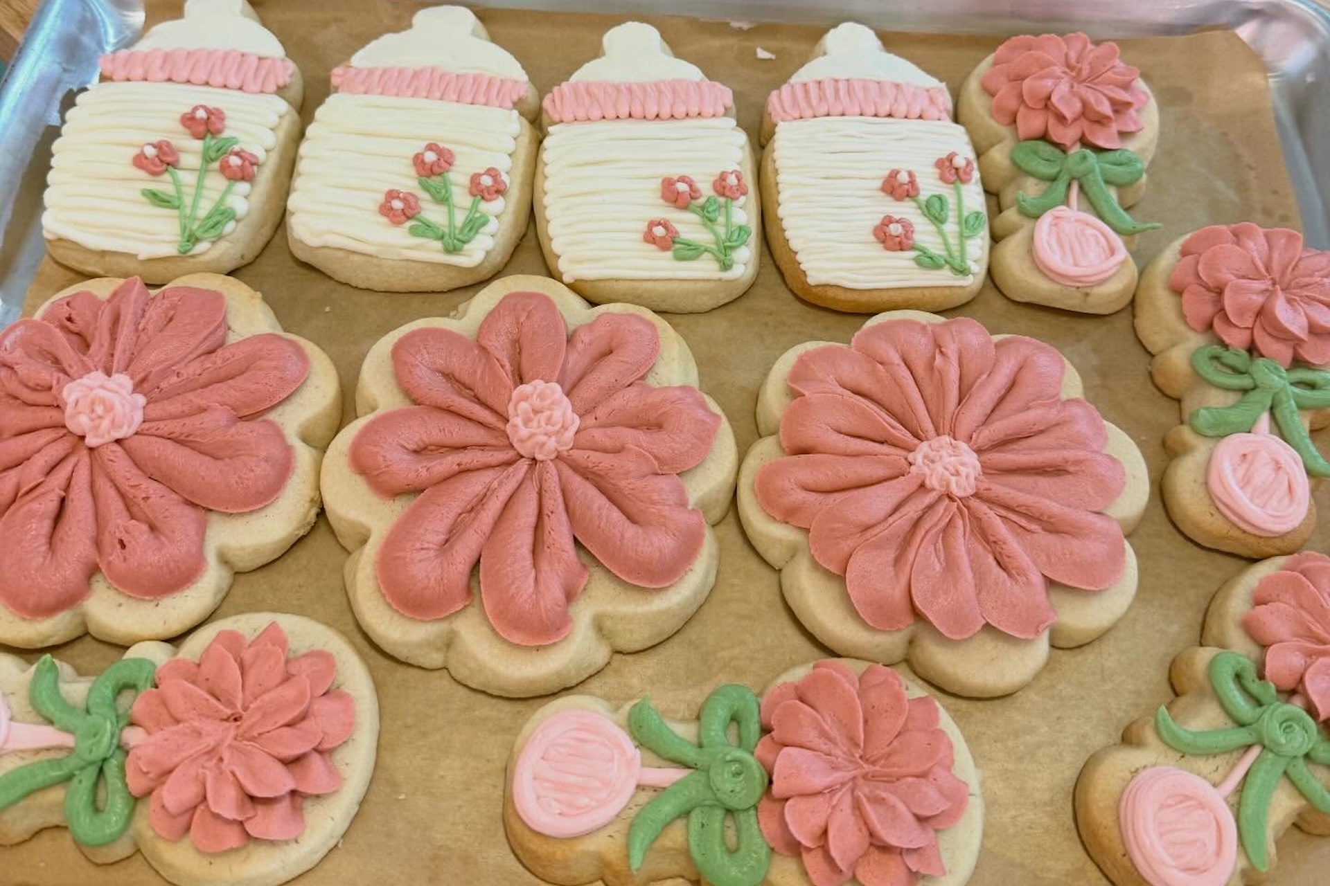 A tray of decorated sugar cookies shaped like baby bottles and flowers, with pink, white, and green icing. Some cookies are large flowers, while others feature small flowers and greenery details.