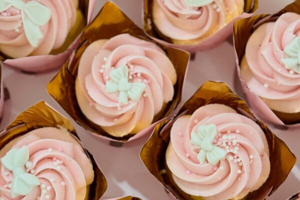 Cupcakes with pink swirled frosting, decorated with white butterfly-shaped toppers and small white sprinkles, are arranged in gold cupcake wrappers.