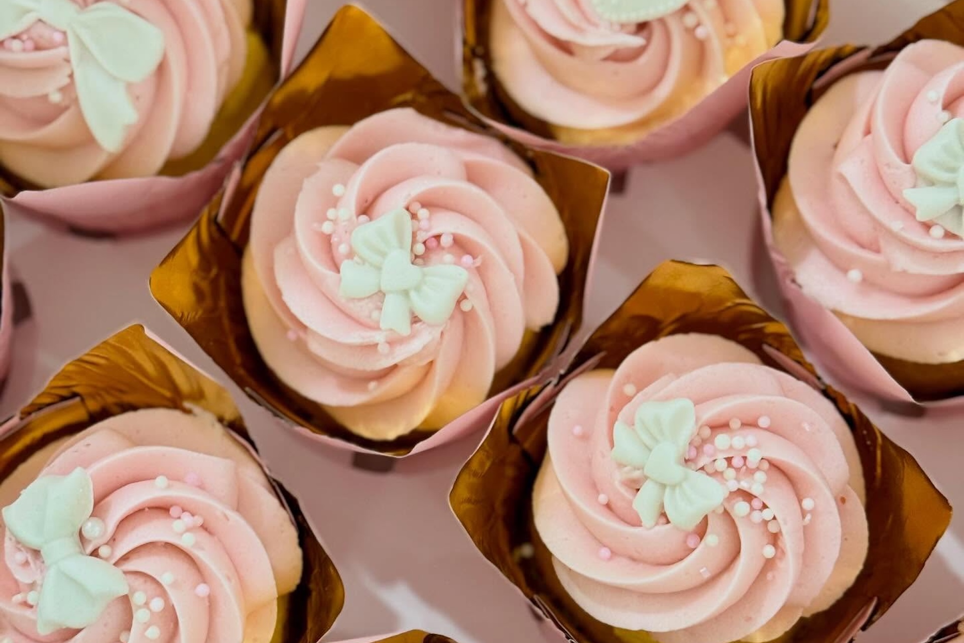 Cupcakes with pink swirled frosting, decorated with white butterfly-shaped toppers and small white sprinkles, are arranged in gold cupcake wrappers.