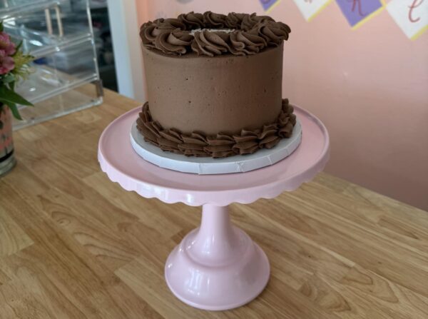 A small, round chocolate cake with chocolate frosting sits on a pink cake stand. The cake has decorative frosting swirls on top and at the base and is placed on a wooden table with a pastel-colored background.