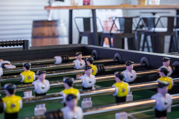 Close-up of a foosball table, focusing on the player figures wearing black and white or yellow and black uniforms, with tables and chairs blurred in the background.