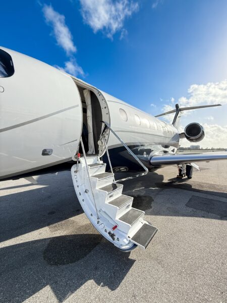 A private jet with its door open and stairs extended sits on an airport tarmac under a bright blue sky with scattered clouds.
