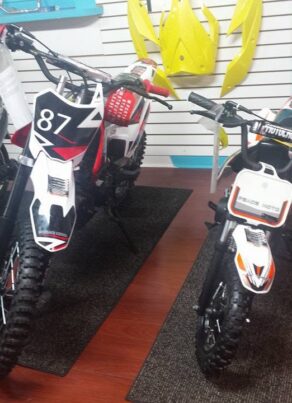 Two off-road dirt bikes, one large and one small, are displayed indoors on dark mats. Both have black and white designs with number plates on the front. Yellow bike parts hang on the wall in the background.