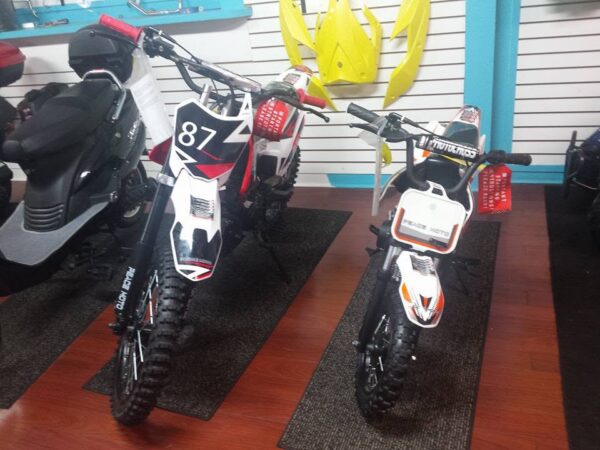 Two off-road dirt bikes, one large and one small, are displayed indoors on dark mats. Both have black and white designs with number plates on the front. Yellow bike parts hang on the wall in the background.