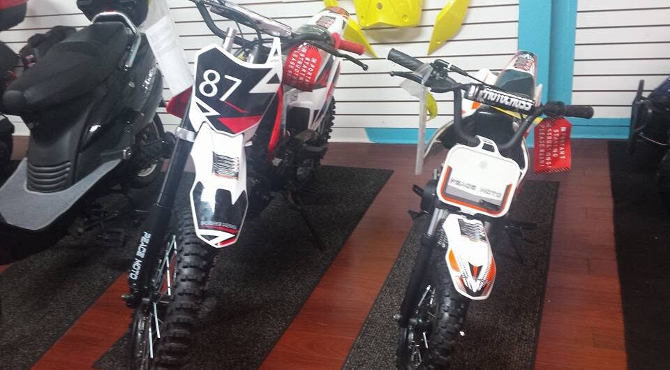 Two off-road dirt bikes, one large and one small, are displayed indoors on dark mats. Both have black and white designs with number plates on the front. Yellow bike parts hang on the wall in the background.