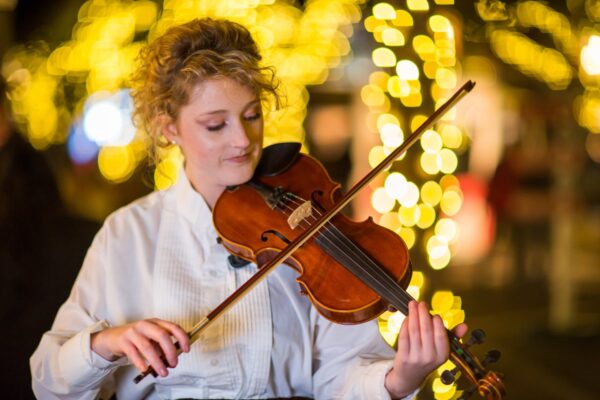A woman with curly blonde hair plays the violin outdoors at night, surrounded by warm, blurred yellow string lights in the background. She wears a white blouse and appears focused on her music.
