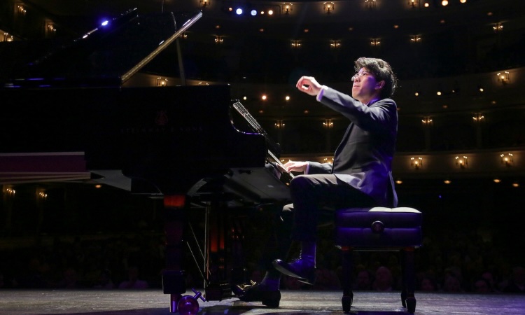 A pianist in a suit playing a grand piano on a stage in a darkened concert hall.