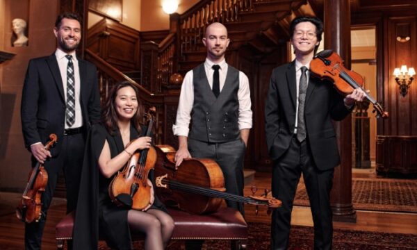 Four musicians in formal attire standing in a wood-paneled room, holding a violin, a viola, a cello, and a violin.