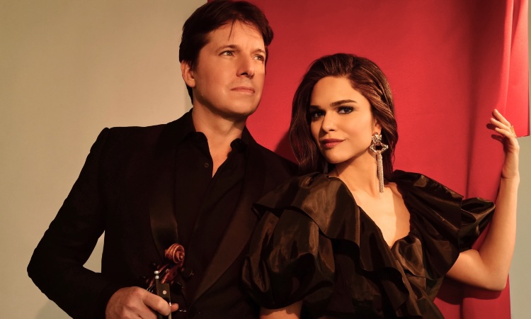 A man in a black suit holding a violin stands next to a woman in a black ruffled dress.