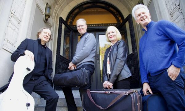 Four people stand in front of an arched building entrance, each holding a musical instrument case.
