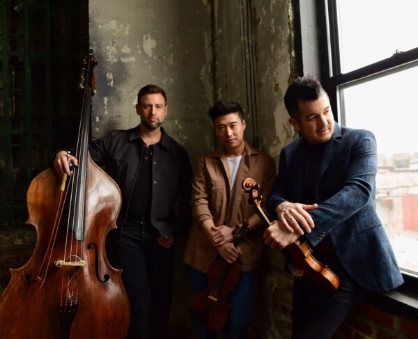 Three men stand by a large window in a rustic room. One holds an upright bass, another a violin, and the third rests a violin on his arm. All look off-camera with serious expressions.