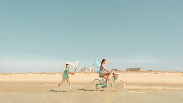 A girl wearing fairy wings rides a bicycle along the beach, pointing forward, while a boy runs behind her holding a butterfly net. Three houses are visible in the background under a clear blue sky.