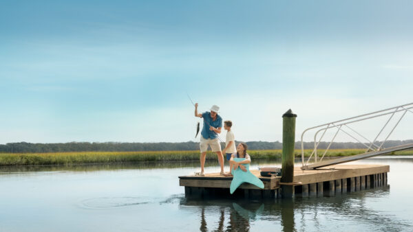 A man and child stand on a wooden dock by calm water, holding up a fish they caught. A woman sits on the edge of the dock, dipping her feet in the water. Grassy marshland and blue sky are in the background.