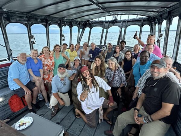 A diverse group of adults smiling and posing together on a boat, some seated and some standing. The boat is covered, with water and a cloudy sky visible in the background. One person is dressed as a pirate.