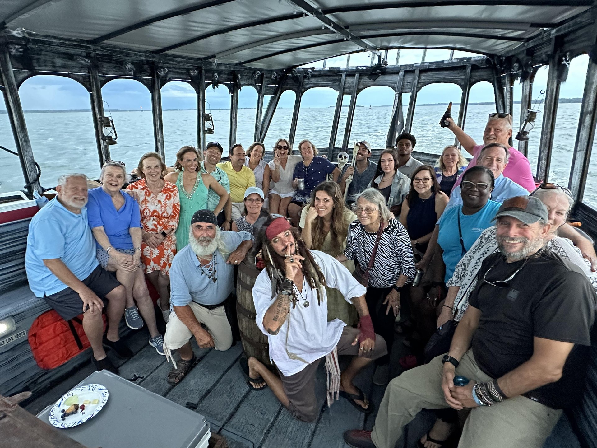 A diverse group of adults smiling and posing together on a boat, some seated and some standing. The boat is covered, with water and a cloudy sky visible in the background. One person is dressed as a pirate.