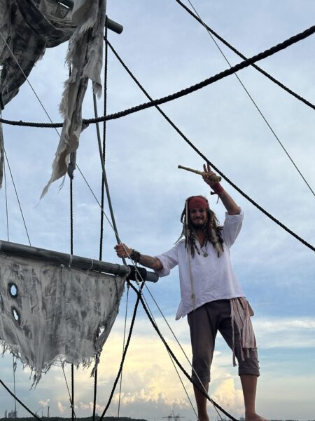 A person dressed as a pirate stands on a ship’s rigging, holding a telescope and smiling. The ship’s sails are tattered, and the sky is partly cloudy in the background.