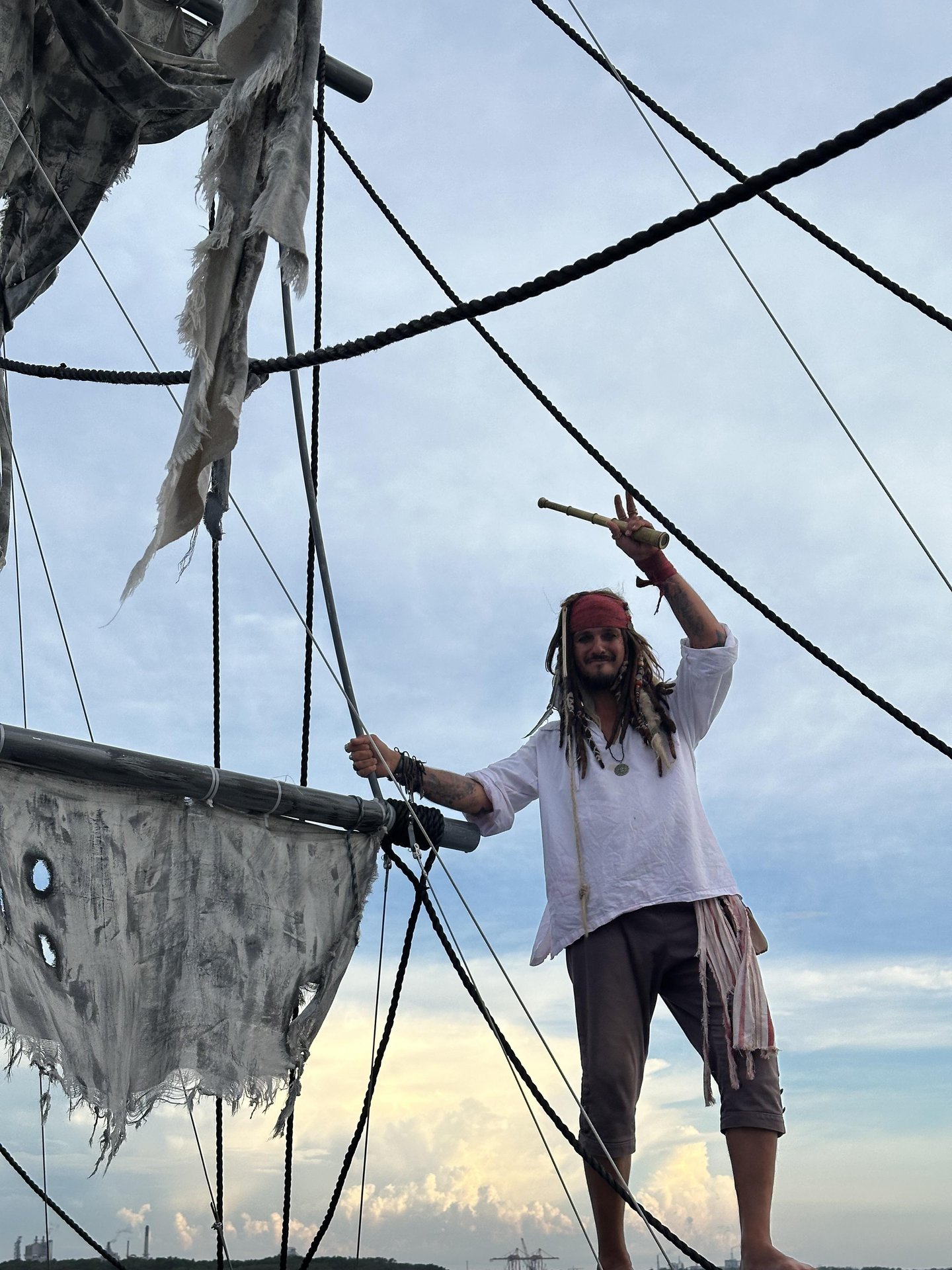 A person dressed as a pirate stands on a ship’s rigging, holding a telescope and smiling. The ship’s sails are tattered, and the sky is partly cloudy in the background.