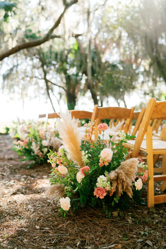 Rows of wooden chairs are set outdoors among trees, decorated with lush floral arrangements of pink and peach flowers, greenery, and pampas grass, creating a serene and elegant wedding ceremony setting.