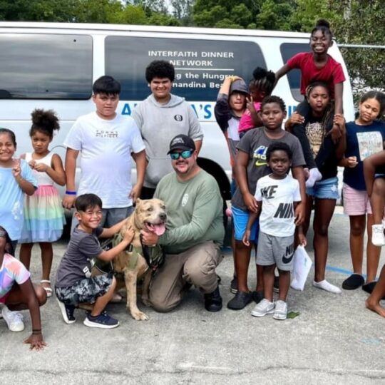 A diverse group of children and adults, along with a dog, pose and smile outside in front of a white van with “Interfaith Dinner Network” written on it.
