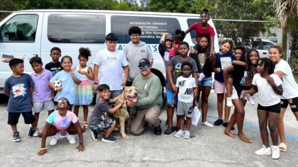 A diverse group of children and adults, along with a dog, pose and smile outside in front of a white van with “Interfaith Dinner Network” written on it.