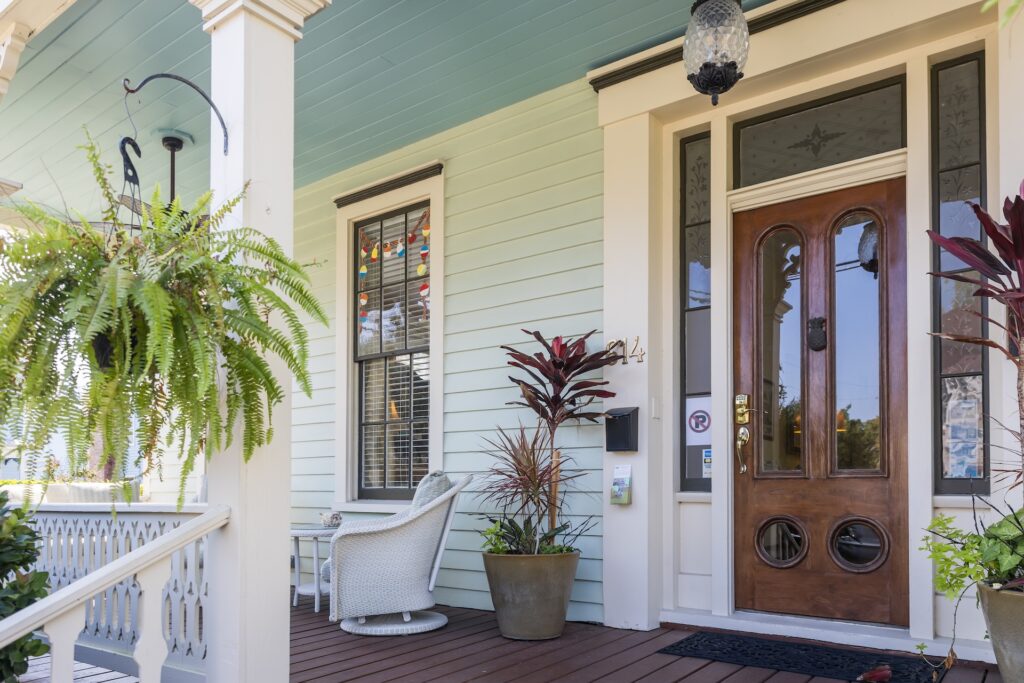 A cozy front porch with a light blue exterior, a wooden door with glass panels, white railing, potted plants, hanging fern, a wicker chair, and string lights reflected in the window.