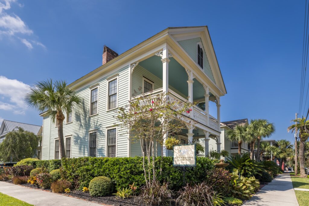 A two-story light blue house with white trim and a wraparound porch, surrounded by lush green landscaping and palm trees, under a clear blue sky. A sign is visible in front of the house near the sidewalk.