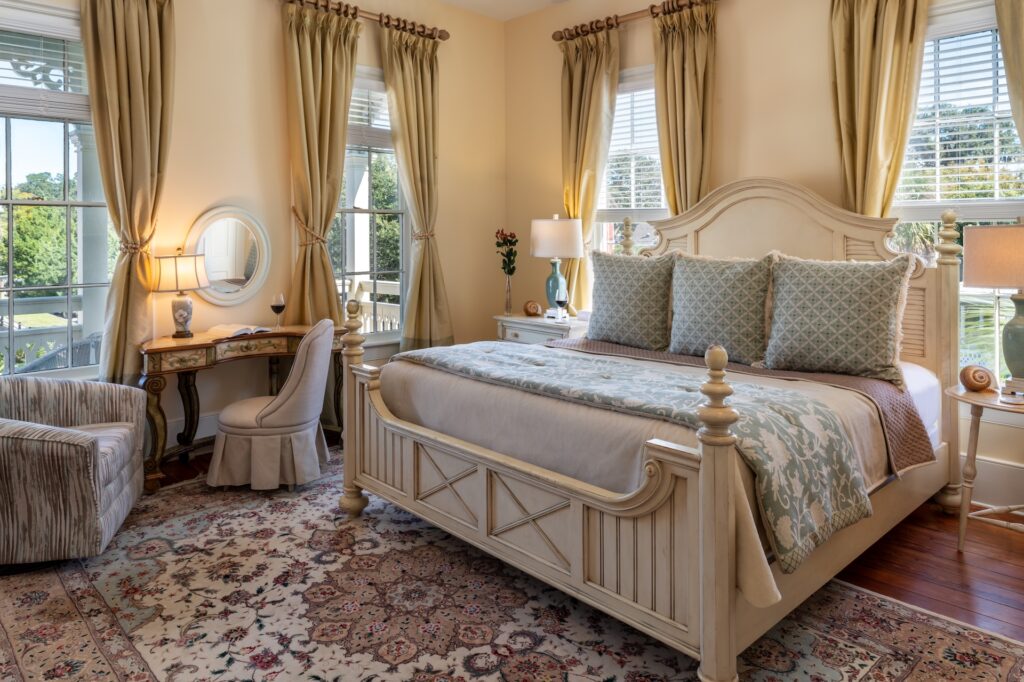 Elegant bedroom with a large bed, patterned pillows and blanket, ornate rug, and classic wooden furniture. Three tall windows with gold curtains let in natural light, and a cozy seating area and vanity complete the space.