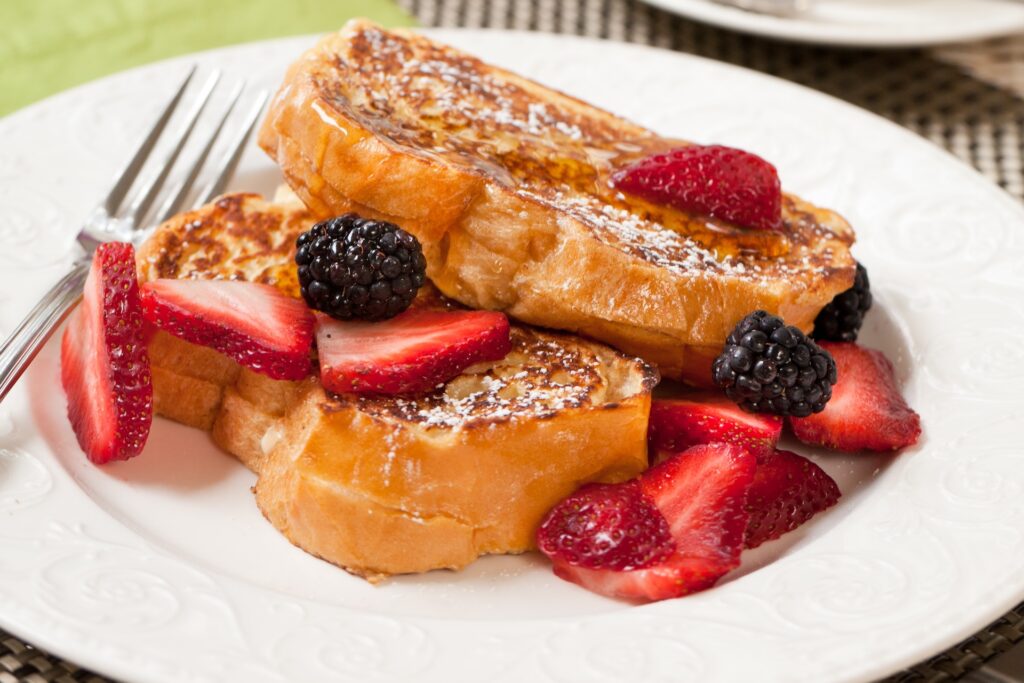 Two slices of French toast topped with powdered sugar, fresh strawberries, and blackberries, served on a white plate with a fork.