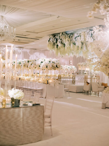 A luxurious wedding reception hall decorated with white flowers, elegant candle centerpieces, and gold accents. White drapery and floral arrangements hang from the ceiling, creating a romantic and sophisticated atmosphere.