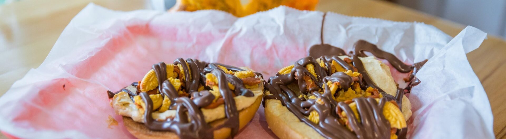 A red basket lined with white parchment holds two bagel halves topped with banana slices, nuts, and drizzled chocolate. In the background, there is autumn decor, including a silver pumpkin and a yellow leaf.