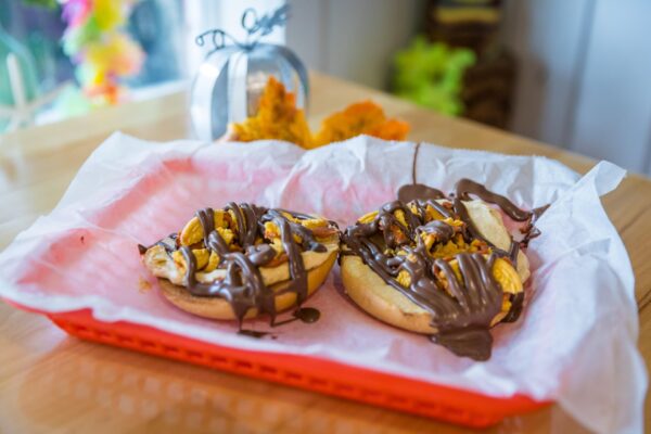 A red basket lined with white parchment holds two bagel halves topped with banana slices, nuts, and drizzled chocolate. In the background, there is autumn decor, including a silver pumpkin and a yellow leaf.