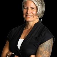 A smiling woman with gray hair and a tattoo on her arm, wearing a black shirt against a black background.