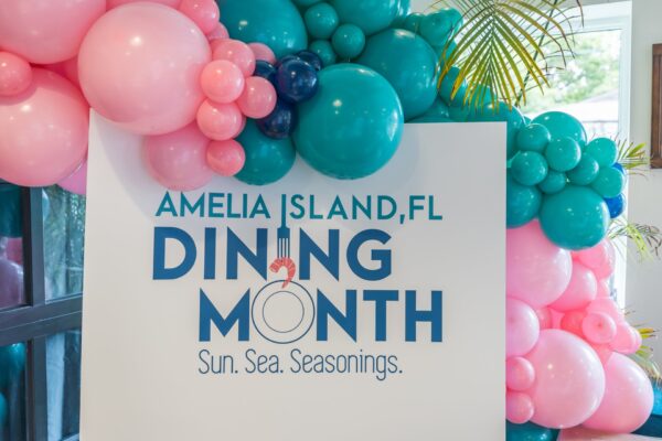 A sign surrounded by clusters of pink, teal, and blue balloons reads, Amelia Island, FL Dining Month. Sun. Sea. Seasonings. Palm leaves and windows are in the background.