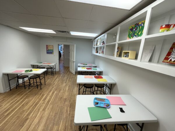 A classroom with white walls and wooden floor has small tables and black stools. Each table has colored paper and art supplies. Shelves with colorful art projects line the wall. A Class Rules poster is by an open door.