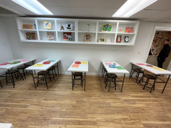 A classroom with white tables and brown stools arranged in rows. Each table has colored paper and art supplies. Artworks are displayed on shelves above and on the wall. A person stands in the doorway to the right.