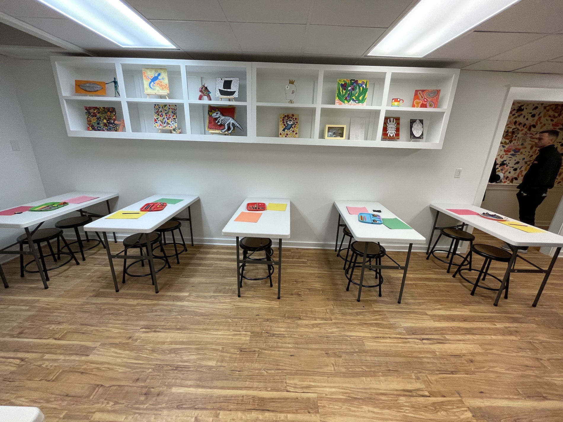 A classroom with white tables and brown stools arranged in rows. Each table has colored paper and art supplies. Artworks are displayed on shelves above and on the wall. A person stands in the doorway to the right.