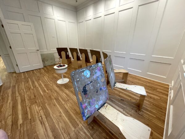 A room with wooden floors, white paneled walls, and benches arranged in rows. The benches have paint splatters, and a small round table with art supplies sits nearby. The door to the room is open.