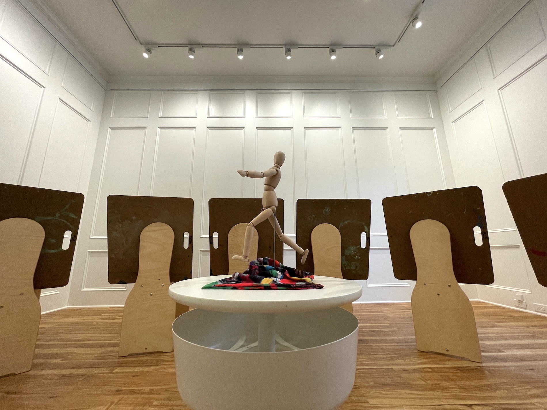 A wooden mannequin stands on a round white table surrounded by abstract wooden cutouts and brown boards in a brightly lit room with wood floors and white panel walls. A colorful cloth lies on the table.