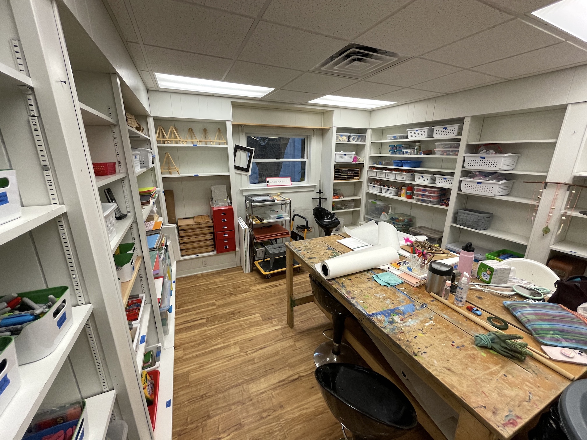 A well-organized craft room with shelves of supplies, a large wooden worktable covered in art materials, storage bins, and a window at the far end. The floor is wood, and the space looks bright and functional.