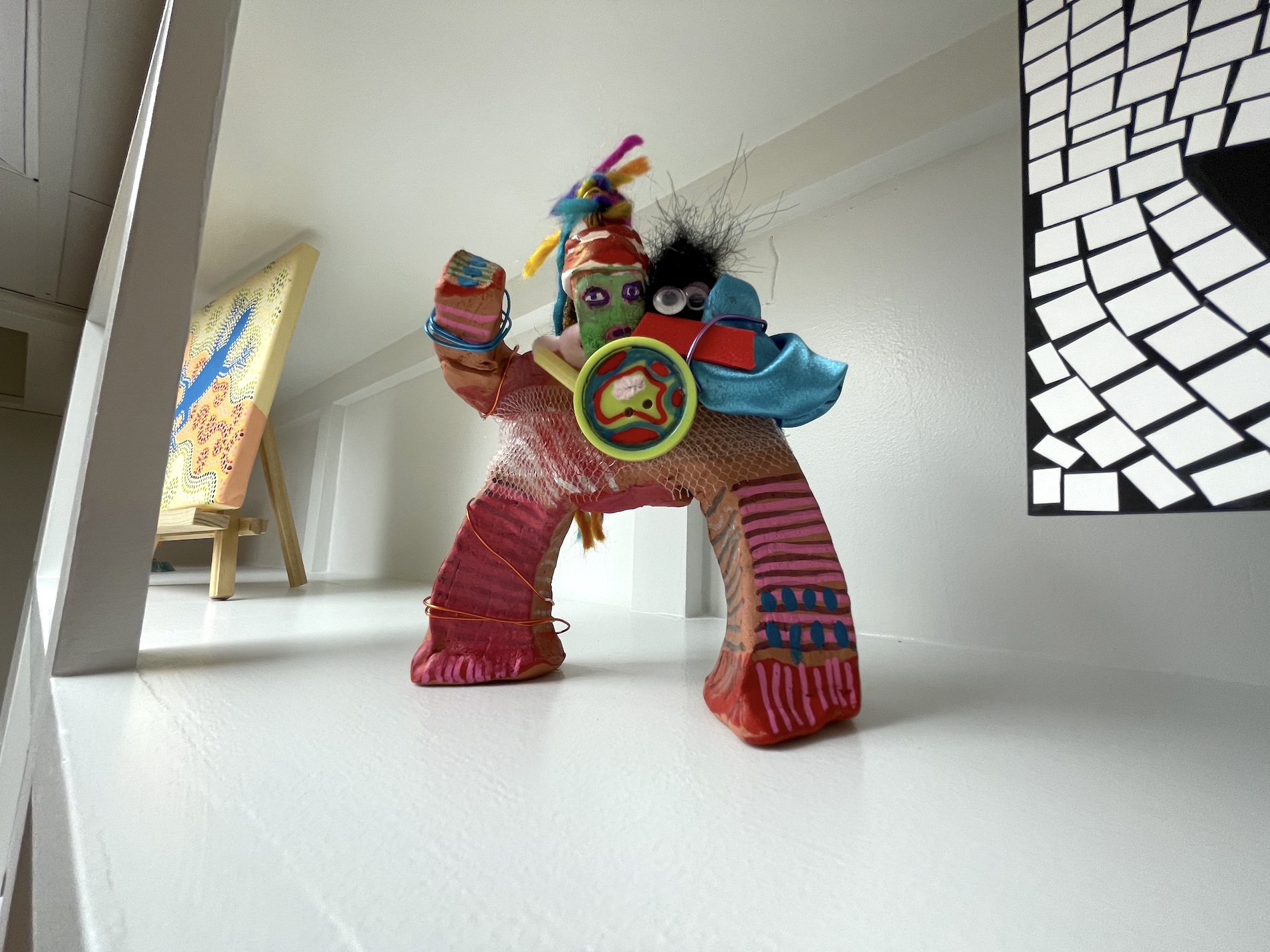 A colorful, handmade figurine with a green face, feathered headdress, and patchwork body stands on a white shelf, surrounded by abstract artwork in the background.